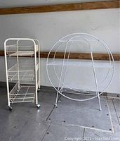 Two metal wire furniture pieces outdoors on a floor with wood rail backdrop; white round wire shelf and cream three-tier rolling rack side by side.