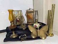 Full view of brass and glass items: amber glass oil lamp, ceramic hand-painted oil lamp, 3D butterfly figure, brass butterfly dish, cylindrical brass sleeves, brass coin organizer, framed mirror piece.