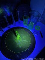 View of the uranium glass pieces glowing under UV light including rosebud vases, cake stand, cruet, and pitcher plus metal pie tin and tools.