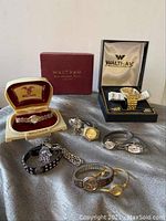Photo showing all 8 watches and 2 bracelets including two Waltham watches in original boxes and a variety of metal bands in gold and silver tones, displayed on a gray cloth background.