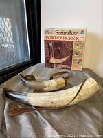 Two curved powder horns placed on a surface next to the vintage Scrimshaw Powder Horn Kit box standing vertically.