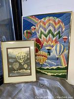 Photo showing large colorful crewel embroidery artwork with multiple hot air balloons in gold frame, and smaller gold foil wall art with matching gold frame, both leaning against a wall on a grey blanket.