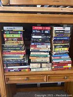 Photo showing a large assortment of paperback and hardcover books stacked in three columns on a wooden desk.