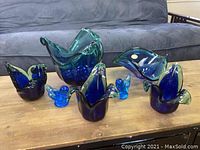 Full view of the six Murano art glass pieces in varying blue shades arranged on a wooden table, showing their shapes and sizes.