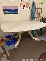 White resin patio table with a rectangular rounded top, shown indoors on a brown carpet. The table has four sturdy curved legs and a hole in the center for an umbrella. Visible surface scratches present.