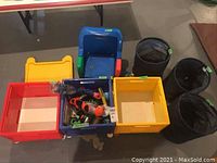 Photo of children's toys including blue high chair, three wheeled storage bins (red, blue, yellow), three black collapsible storage bins, and toys inside bins on a gray floor.