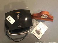 View of George Foreman panini grill, wooden mantel clock, and instruction manual on floor.