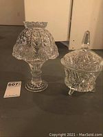 Two crystal footed bowls with lids placed side by side. One tall with a scalloped rim and one shorter with a finial lid and three feet.