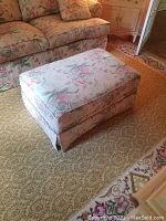 Front angled view of large rectangular ottoman covered in floral fabric upholstery with pink and green tones, matching a nearby sofa, placed on patterned carpet.