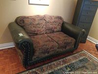 Overstuffed loveseat with floral cushions and dark green leather armrests, slightly worn on the arms, placed near a blue cabinet and a rug on a wood floor.