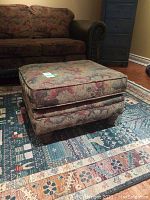Front angled view of a fabric-upholstered ottoman with vintage-style multicolor floral design on the fabric and wooden legs on a patterned rug near a couch.