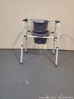 Front view of the portable commode with adjustable legs, plastic bucket inserted, and a lid hanging on the frame.