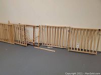 Five wooden baby gates arranged side by side against wall, one gate is visibly cracked, some hardware pieces on floor.