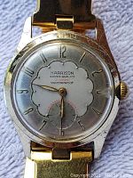 Close-up of the watch face showing brand Harrison, 21 jewels, and waterproof marking with gold-tone numerals and hands, showing scratches on crystal and wear on case.