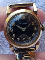 Close-up view of ALPA wristwatch with black dial, gold-tone case, and silver-tone expandable band showing details of the dial.