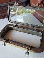 Porthole with open hinged glass showing intact gasket and brass frame with wing nut latches.