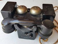 Front and top view showing handset in cradle, dual brass bells, and black heavy-duty body.