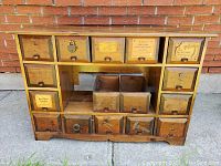 Front view of antique cabinet showing 15 wood and tin drawers with various vintage labels and metal handles, in front of brick wall.