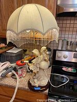 Side view of bisque fairy figurine table lamp with cream-colored fabric shade and beaded fringe. Lamp is lit showing functionality.