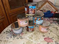Overall view of six stacked candles in glass jars with metal lids on table showing various scent labels.