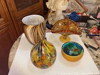 Photo showing all four pieces together: brown and white striped vase, bulbous multicolored vase, multicolored bowl with yellow outside, and amber fan-shaped piece on pedestal.