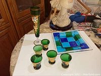 Photo showing the tall green decanter jug with gold and floral hand painted details, along with five matching small green glasses and a square blue and green glass tray.