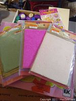Photo showing three packs of textured paper in green, pink, and cream, placed on top of a box containing other crafting supplies.