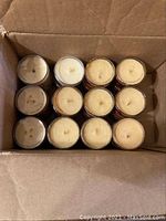 Top view of 12 yellowish cream wax candles in glass jars inside a cardboard box. Used condition with some discoloration on wicks.