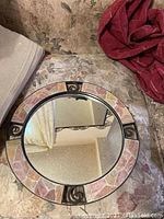 Round mirror with mosaic stone frame and metal scroll details, placed on patterned fabric.