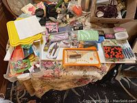 Wide view of table with mini paper cutter, letter stickers, various threads, cords, glitter, and assorted sewing and crafting supplies scattered on tablecloth.