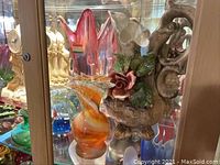 Photo showing a large ornate ceramic Capodimonte pitcher with floral decorations, a pink and white Murano glass vase with orange swirl in front, and other glass items in background.