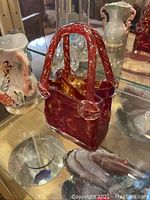 Red marbled Murano glass purse with clear handles placed on glass shelf.
