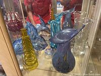 Four decorative Murano and Venetian glass pieces displayed in cabinet, showing colors and shapes including blue flower, purple vase, yellow vase, and clear glass vase.