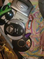 Photo showing several portable music players including a silver Sony Walkman cassette player, a black Sanyo Discman portable CD player, a red and silver MP3 player, along with various USB plugs, a white power adapter, and black headphones all set on a fabric background.