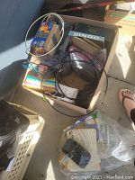 Box with assorted small items including bingo game, cables, party blower, and alarm clock.