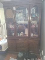 Full front view of the dark wooden china cabinet showing the three glass doors with metal lattice design, lower storage cabinets and drawers, visible damage to front glass, and missing glass shelves. The room and some contents inside the cabinet are visible but not included.