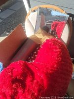 Photo showing a red heart-shaped pillow, brown cardboard box, dark cylindrical object (vase), and a rectangular mirror with a wooden frame placed on a cushioned chair.