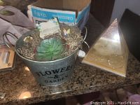 Metal bucket with faux succulent plant labeled 'FLOWERS & GARDEN' on a counter