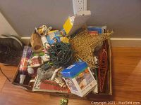 Box of assorted holiday and kitchen items including Christmas village pieces, strand lights, star decoration, cake pans, and bottle opener.