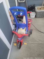 Rear view of red and cream toddler push trike with blue tall push handle on concrete floor