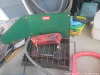 Photo showing green metal camping stove with two burners and a red fuel tank attached, placed on a concrete surface with garden hose and other storage items in background.