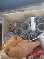 Photo showing cardboard box with multiple glass jars and containers holding assorted nails and screws, along with a partially visible brown paper bag.