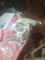 Close-up photo of a hand holding a small plastic bag containing various pieces of costume jewelry including braided bracelets with wooden beads and metal charms in assorted colors.