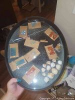 Photo showing a hand holding a black glittery oval tray with multiple resin jewelry pieces in plastic sleeves on kraft paper backing, plus several off-white spherical objects clustered on one side.