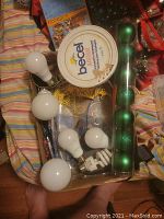 Box containing several white incandescent and CFL light bulbs, green Christmas ornaments in clear tube, Becel tub, and items including light bulbs.