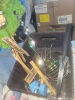 Brown plastic basket holding several gardening and kitchen utensils including metal berry picker, twisted ladle, whisk, wooden spoon, wooden clothespins clipped to a pink cloth, and a black plastic garden dibbler, among other tools.