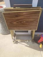 Image showing four wood-grain TV trays stacked vertically on a metal wheeled rack against a wall, highlighting the tray surfaces and rack base.