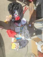 Photo showing lot items including two children's backpacks, stuffed teddy bear, miniature hockey sticks, children's books, and various toys and clothing items laid out on a concrete area.