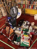 Wide view showing tennis racquet, walking sticks, golf clubs, golf balls, gloves, weights, flags, mugs, and other miscellaneous items on red carpet floor with wood paneled background.