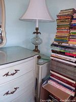 Tall white wooden side table with glass top and bronze decorative hardware beside a stack of books.